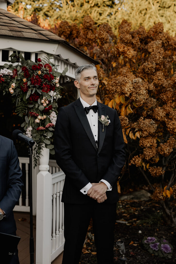Groom smiling at the gazebo with fall foliage behind at The Farmhouse wedding venue in Hampton, NJ while he watches the bride walk down the aisle.