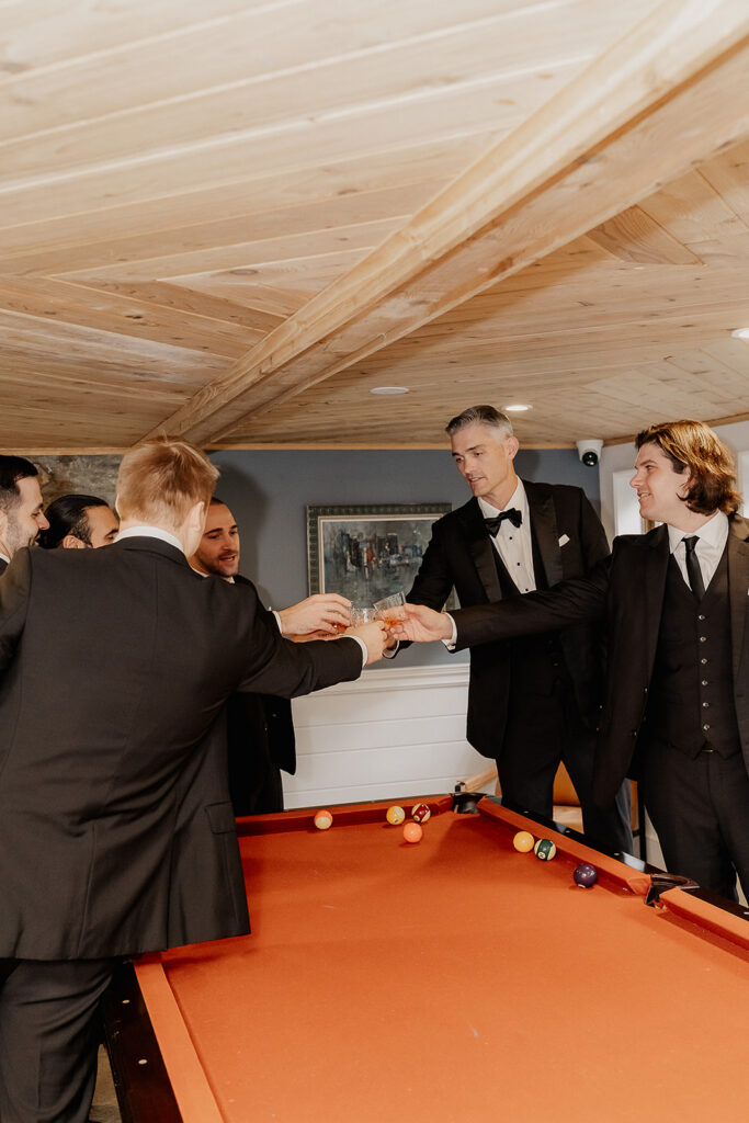 Groomsmen cheering around a pool table during getting ready at a farmhouse wedding in Hampton, NJ.