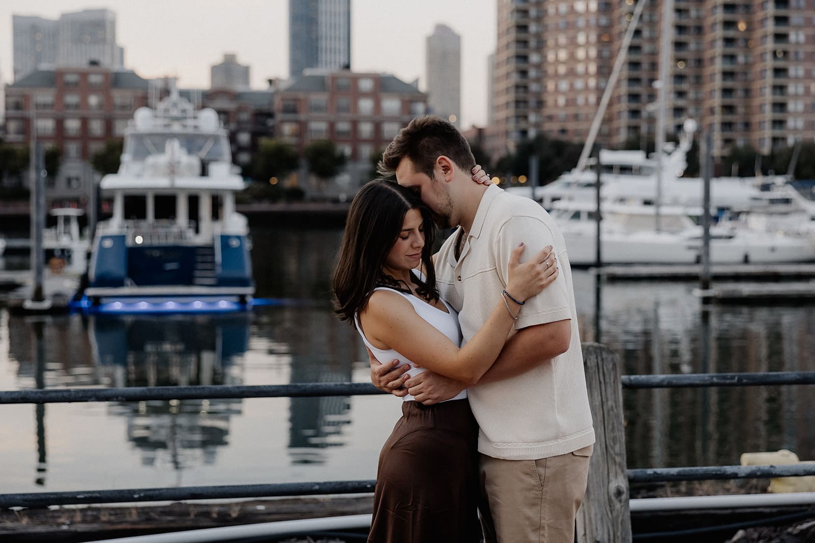 Couple posing in front of boats in the water and buildings for their Liberty State Park engagement photos in Jersey City, NJ.