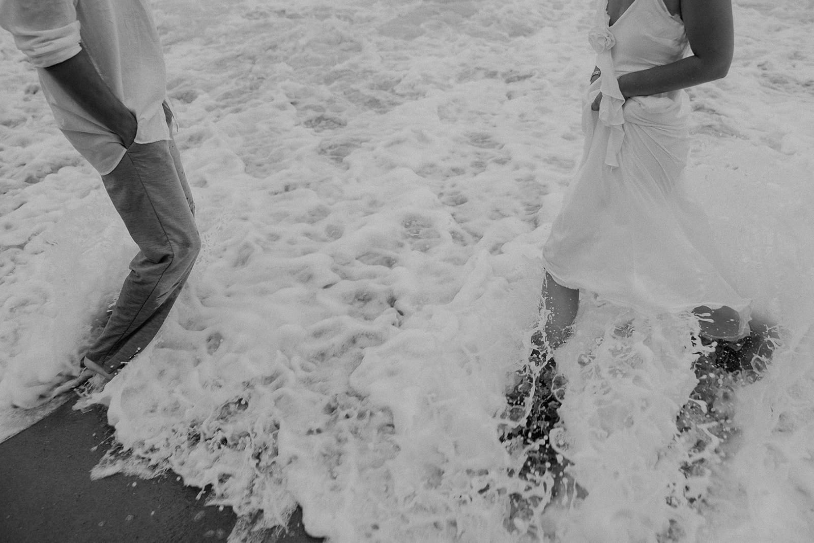 Black and white photo of waves crashing into a couple who are standing in the water.