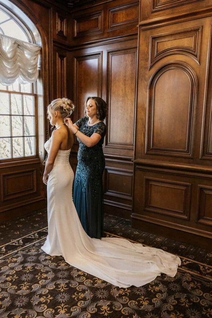 Brides mother adjusting her necklace by a window at Brides dress hanging on the wall at The Brownstone in Paterson, NJ.
