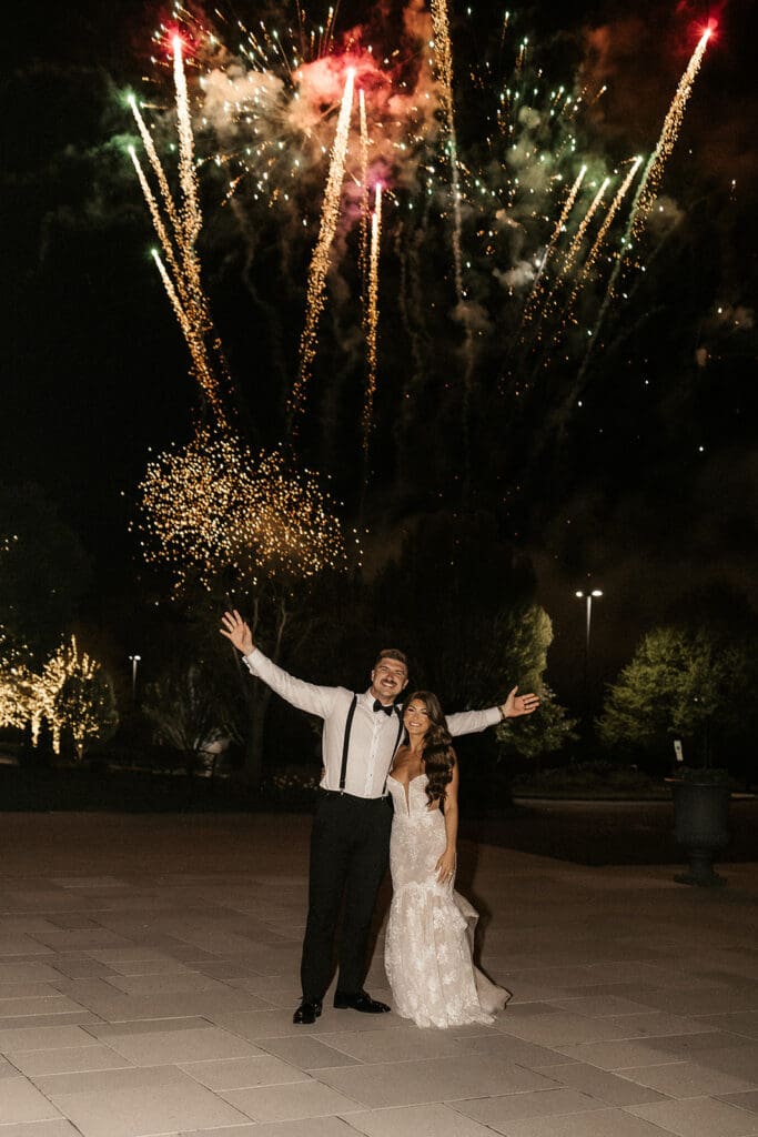 Bride and groom posing outdoors while fireworks shoot off behind them during their wedding reception.