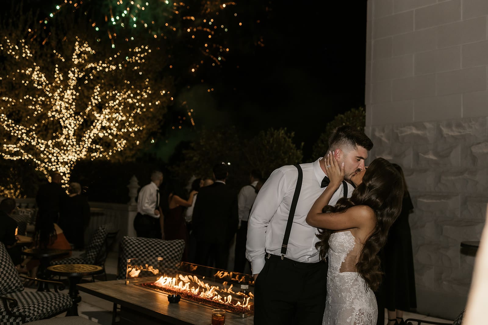 Bride and groom posing outdoors while fireworks shoot off behind them during their wedding reception.