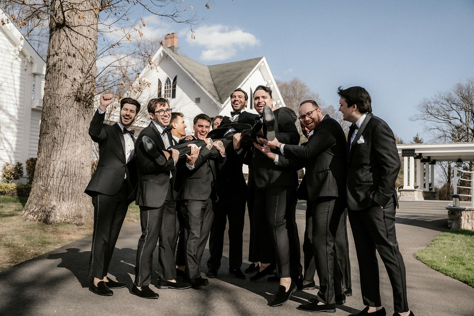Groomsmen in black tuxedos lifting the groom up during bridal party photos at The Ryland Inn.