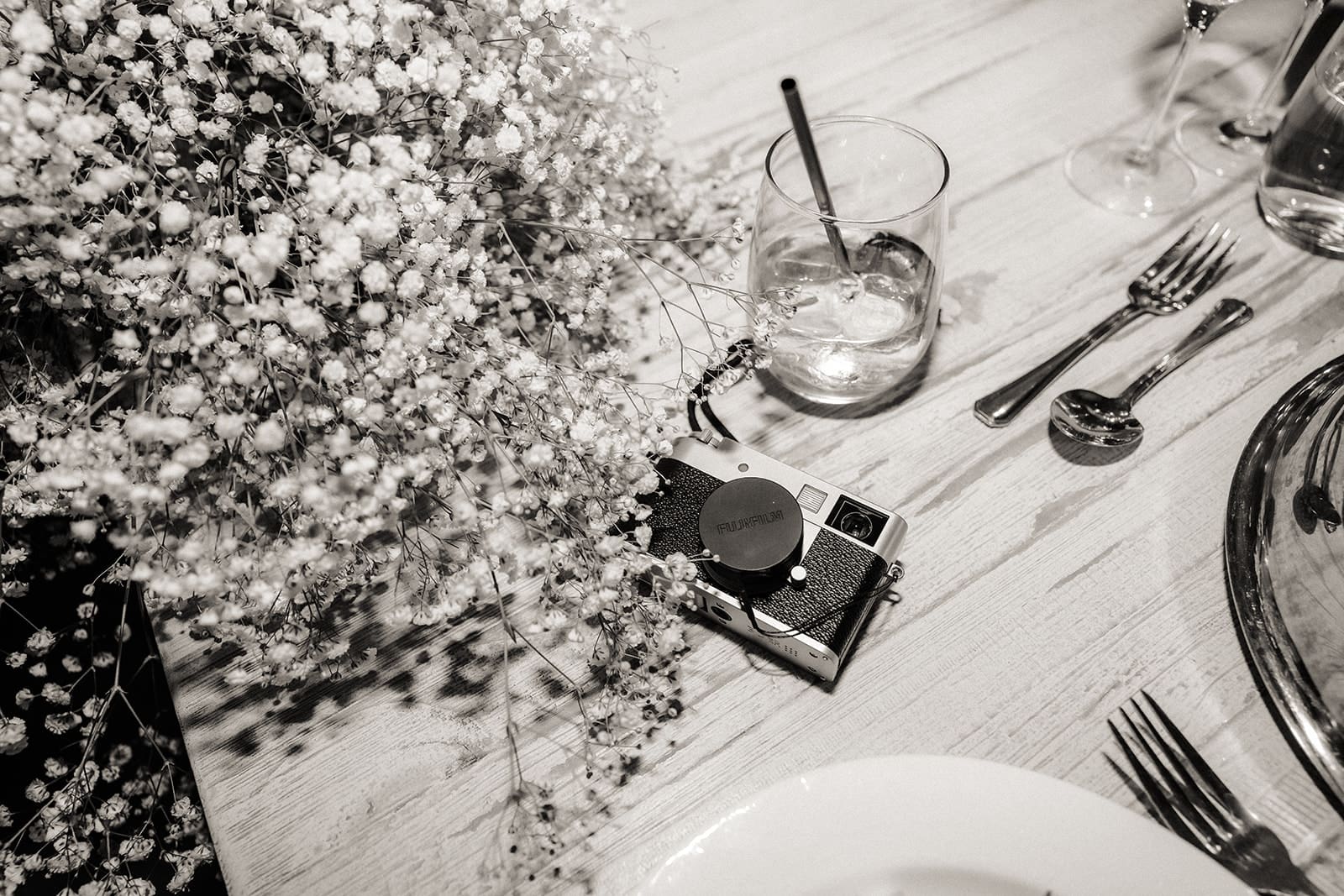 Black and white wide shot of wedding reception details with a fuji camera, half drunken drink, and florals.