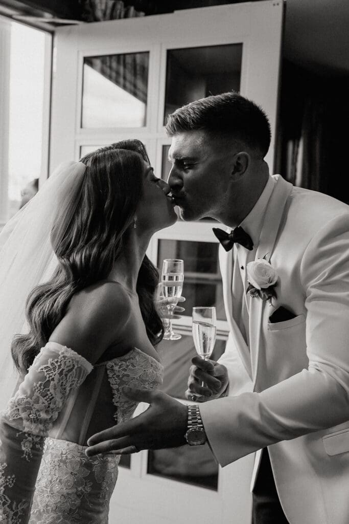 Black and white photo of a bride and groom kissing with champagne after their ceremony.